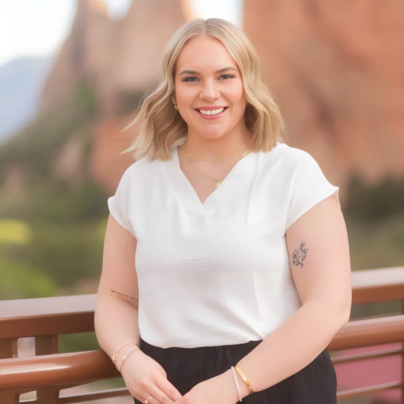 Smiling woman in white blouse standing outdoors with mountains in the background.