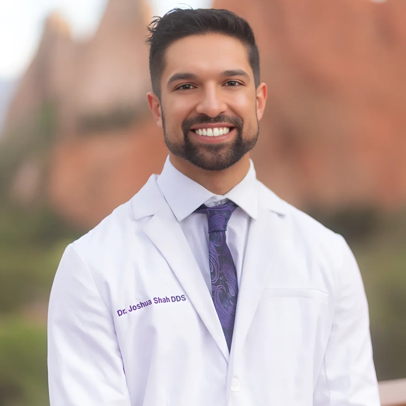 Smiling male doctor in white coat with blurred outdoor background.