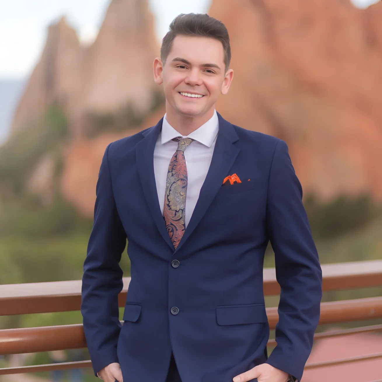 A man in a navy blue suit smiling outdoors with rocky formations behind.