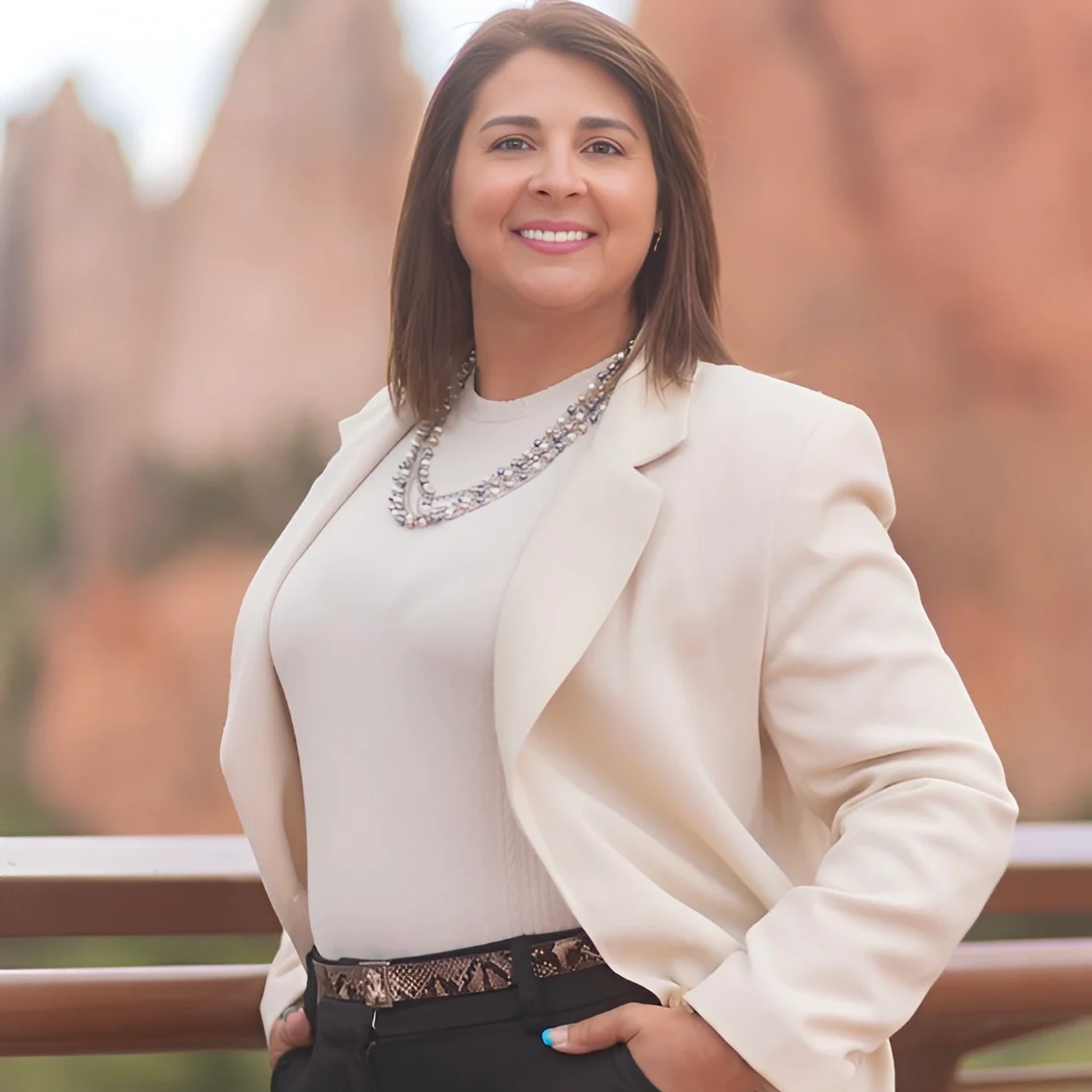Smiling woman in a beige blazer outdoors with natural rock formations in the background.